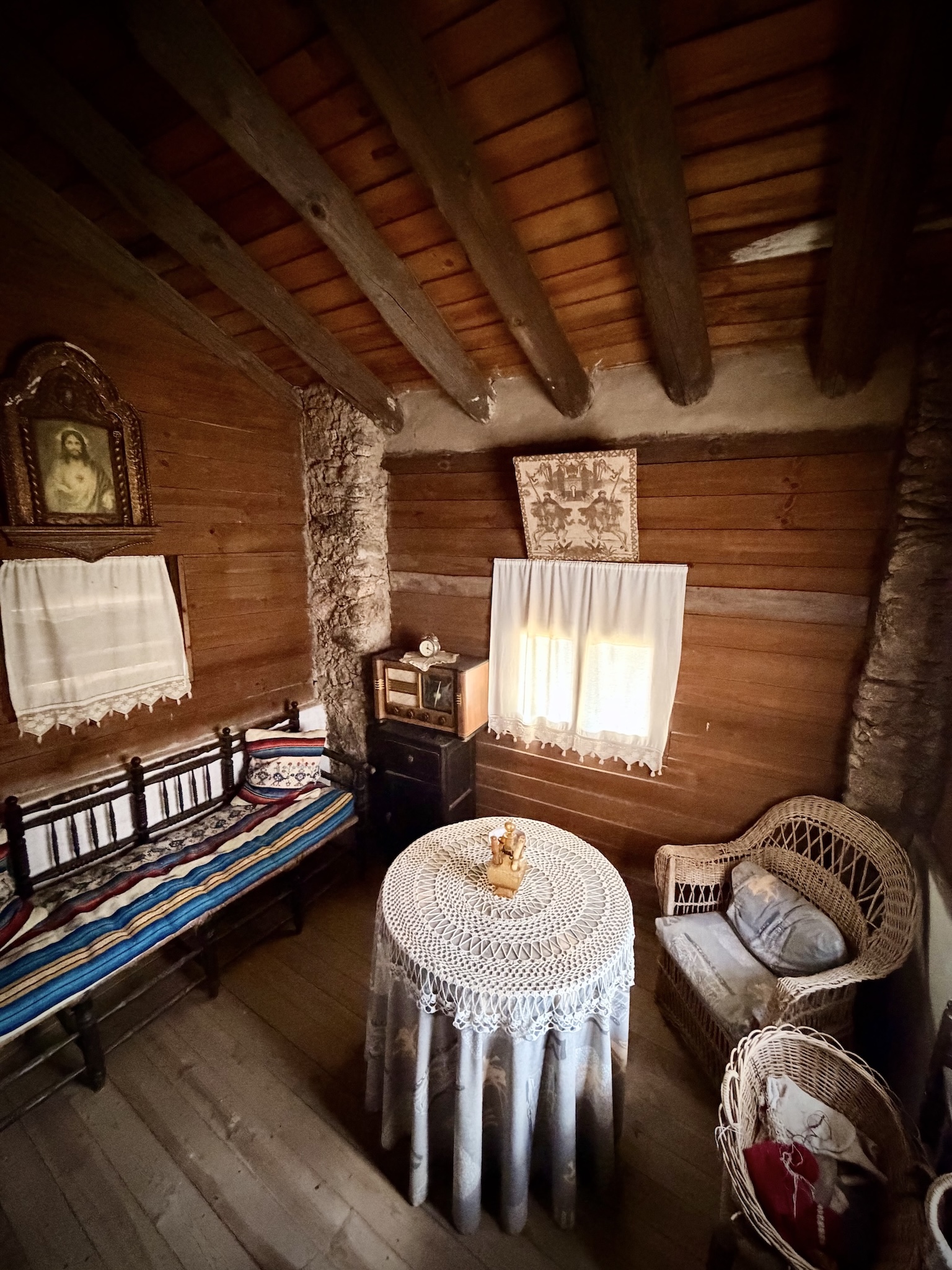 Rinconcito cozy en una parte del museo etnográfico que visité. Hay una mesa camilla con un mantel de ganchillo, un sillón de mimbre, otro de madera, una radio antigua y un tapiz.